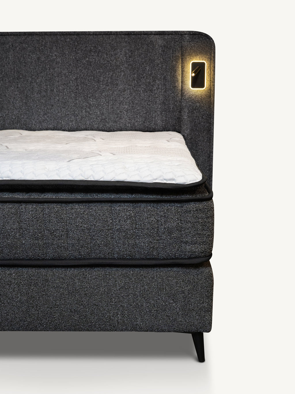 A close-up of a dark gray upholstered bed frame with a white mattress topper and a built-in warm light on the headboard, shown against a plain light background.