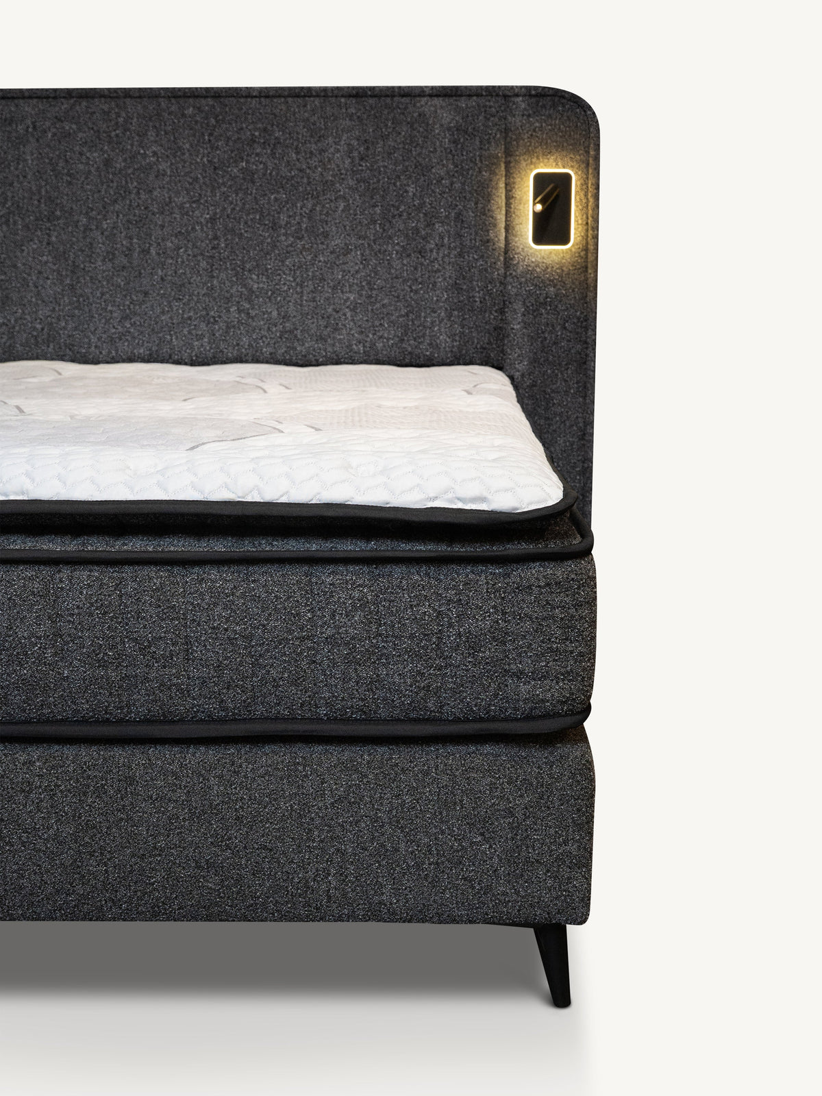A close-up of a dark gray upholstered bed frame with a white mattress topper and a built-in warm light on the headboard, shown against a plain light background.