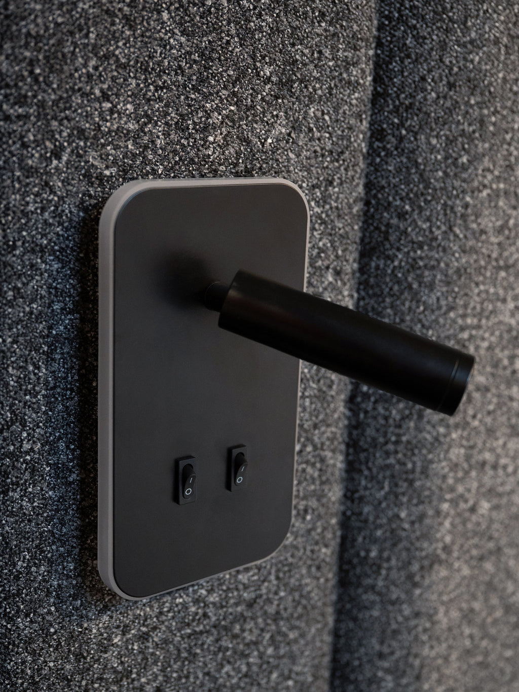 A close-up of a modern matte black wall-mounted power strip with two switches, set against a textured dark gray wall.