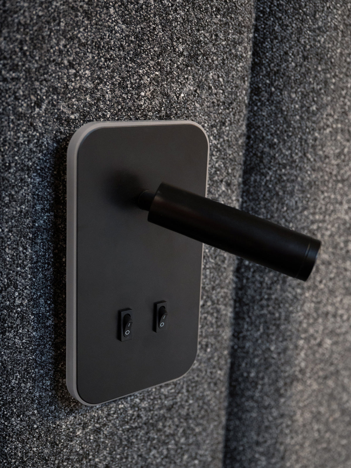 A close-up of a modern matte black wall-mounted power strip with two switches, set against a textured dark gray wall.