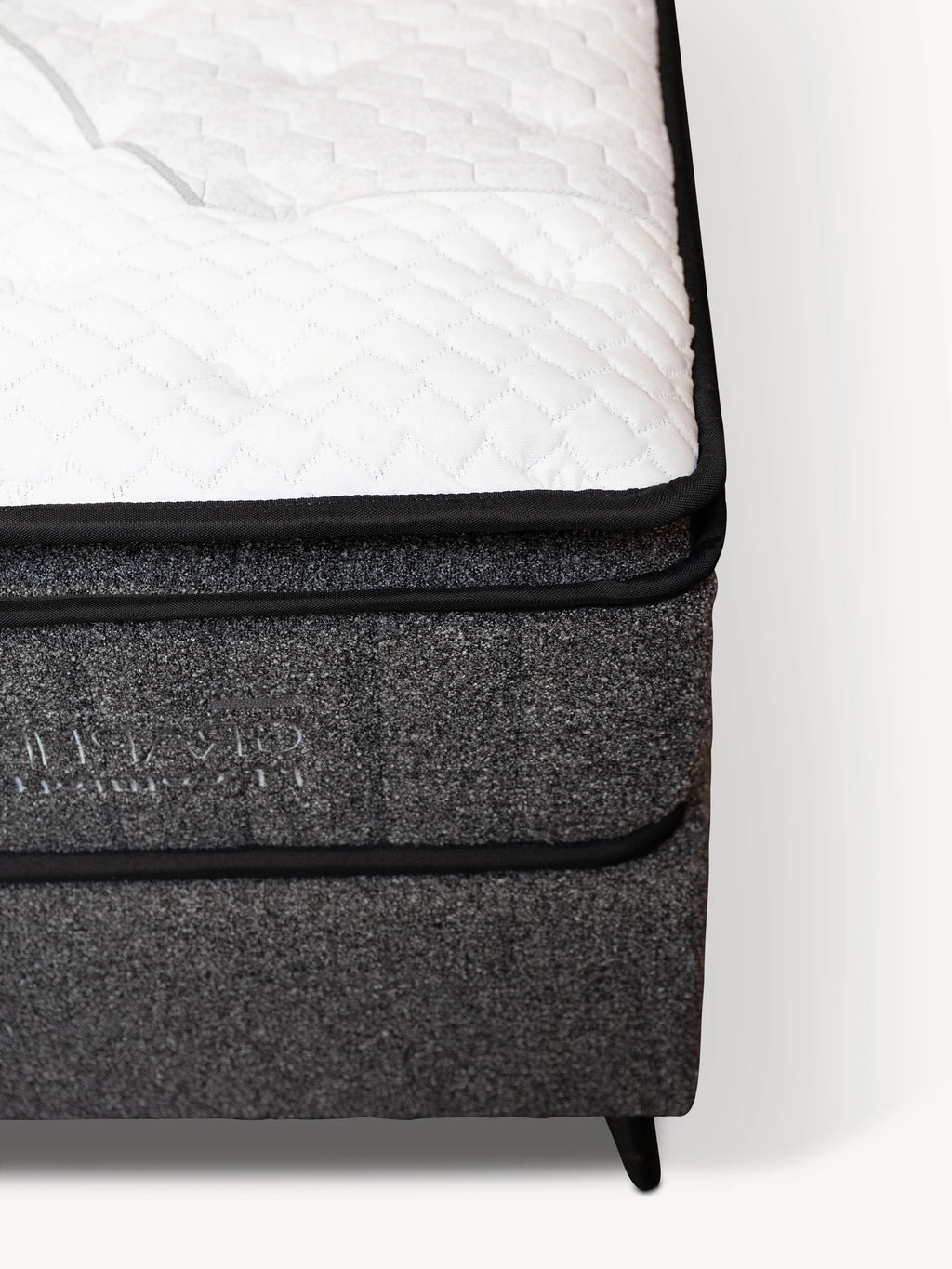 Close-up of a mattress corner showing a white quilted top surface, black piping, and a dark gray textured side panel with debossed branding.