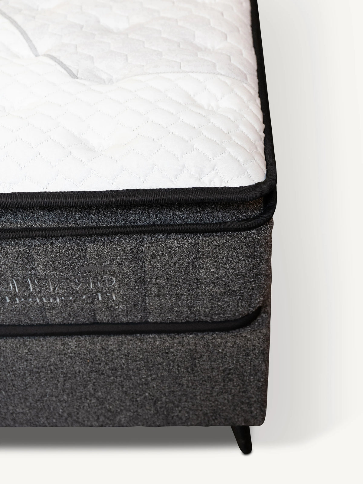 Close-up of a mattress corner showing a white quilted top surface, black piping, and a dark gray textured side panel with debossed branding.