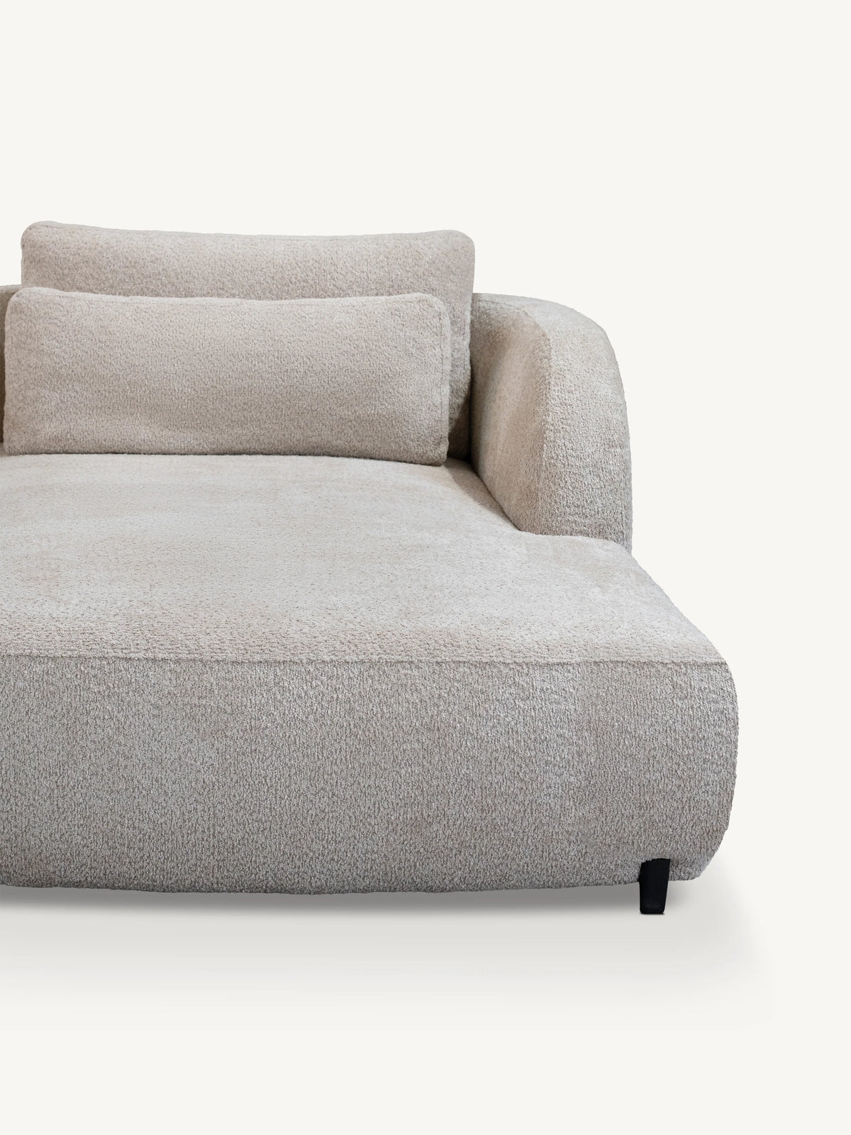 A close-up of a modern chaise lounge chair upholstered in a textured bouclé fabric in a light beige-gray color, featuring a low-profile seat, a wide armrest, and two matching back cushions.