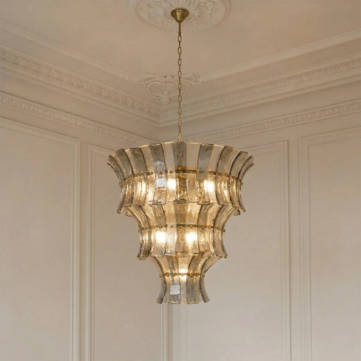 A tiered chandelier with fluted clear glass panels and warm brass-toned metal hardware, hanging from a chain in an elegant cream-colored room with decorative wall molding and a coffered ceiling.