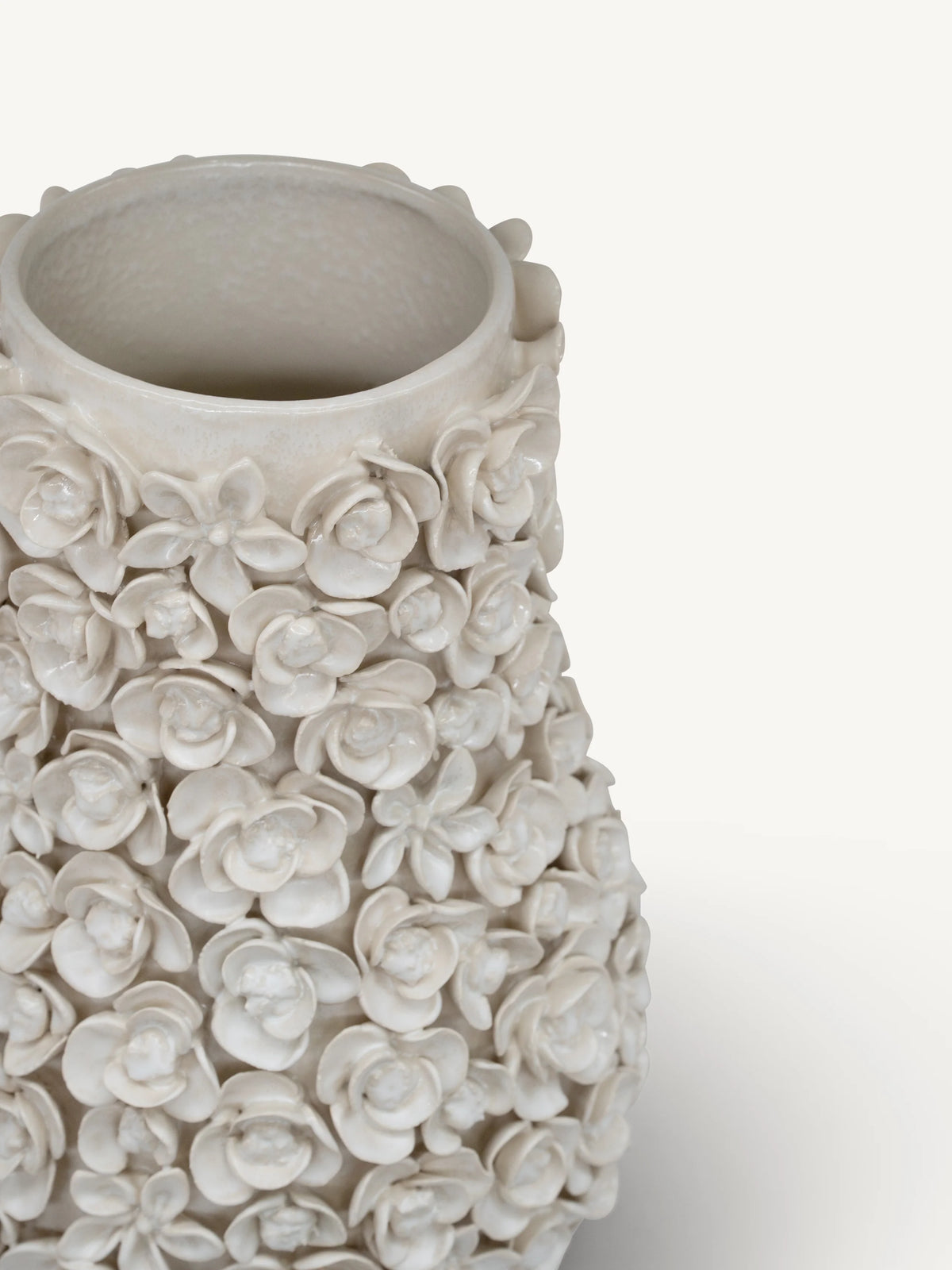 A close-up of a textured ceramic vase with a dense, 3D floral relief pattern in a warm off-white or cream glaze, set against a plain light background.